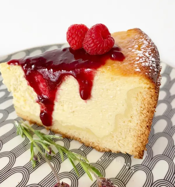 Ein saftiger Käsekuchen mit roter Fruchtsauce und frischen Himbeeren wird auf einem dekorativen Teller präsentiert.