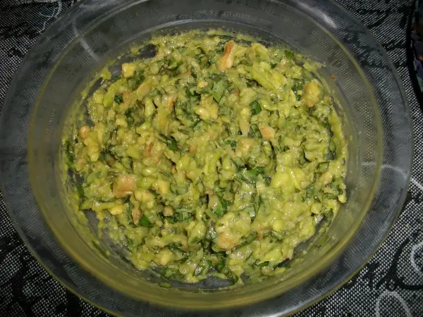 Eine grüne Mischung aus Avocado, Basilikum und Walnüssen in einer Glasschüssel steht auf einem dunklen Tisch.