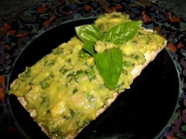Ein knuspriges Brotstück mit Avocado und Basilikum liegt auf einem schwarzen Teller, umgeben von einer bunten Dekoration.