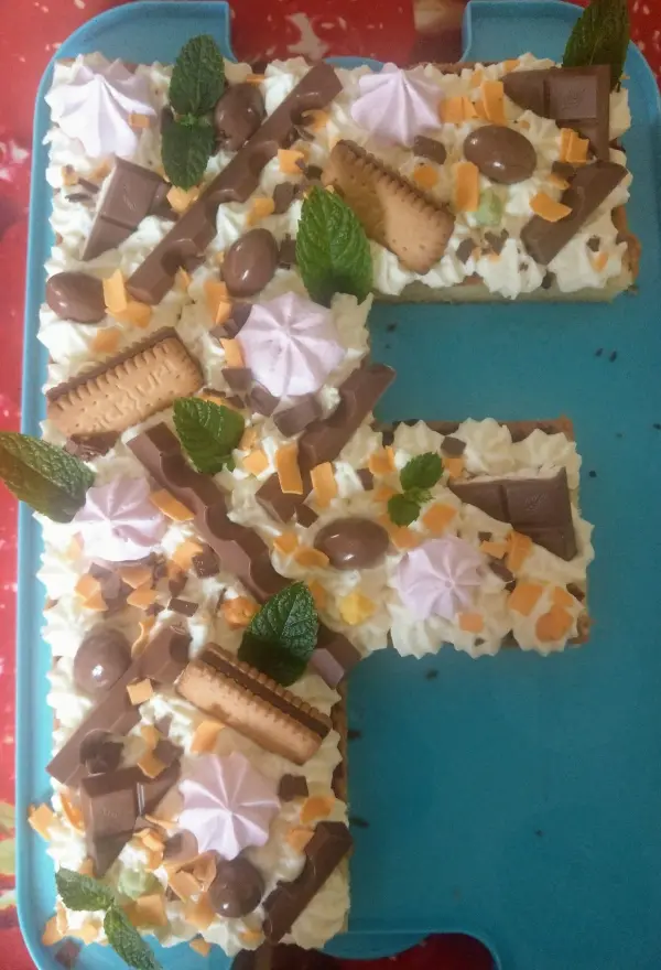 Ein bunter Number Cake in Form der Zahl E, dekoriert mit Süßigkeiten und frischen Minzblättern, präsentiert auf einer blauen Platte.