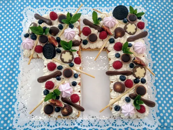 Ein dekorativer Number Cake in Form der Zahl 17 liegt auf einem blauen Tisch mit weißen Punkten, umgeben von frischen Früchten und Süßigkeiten.
