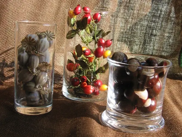 Glasbehälter mit dekorativen herbstlichen Elementen wie Beeren und Samen stehen auf einem braunen Stoffhintergrund.