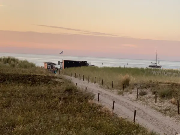 Ein Weg führt durch das Gras zu einem Strand in Warnemünde, wo ein Boot vor einem sanften Sonnenuntergang ankert.