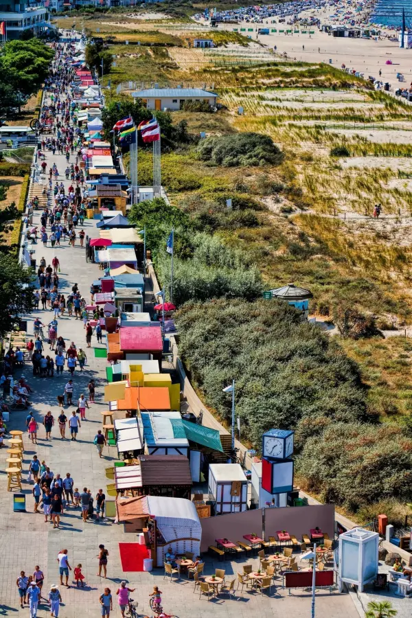 Bunte Verkaufsstände entlang einer belebten Promenade in Warnemünde, umgeben von Sanddünen und erholenden Besuchern.