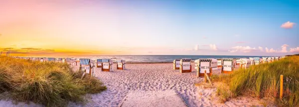Weißer Sandstrand mit liegenden Strandkörben, während die Sonne hinter dem Horizont aufgeht, an der Ostsee in Warnemünde.