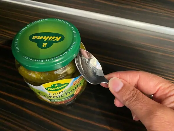Ein Teelöffel öffnet den Deckel eines Schraubglases auf einer Holzoberfläche, um den Inhalt zugänglich zu machen.