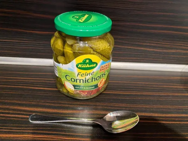 Ein Schraubglas mit Gurken steht auf einer Holzfläche, daneben liegt ein Silberlöffel, der zur Entnahme dient.