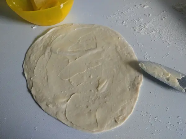 Ein Laugendreieck wird vorbereitet, indem ein glatter Teig ausgewalzt und mit einer Kelle bestrichen wird.