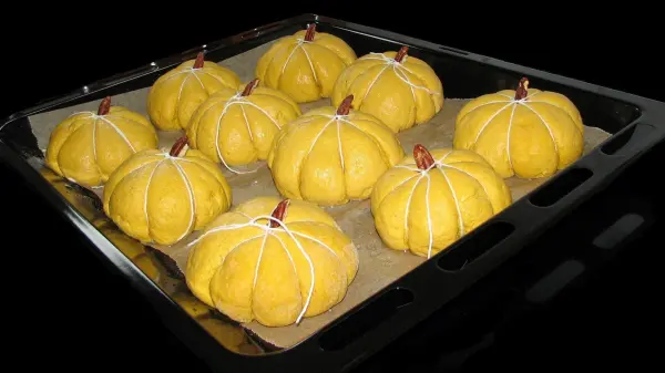 Auf einem Backblech liegen mehrere Kürbisbrötchen, die rund und gelb sind, bereit zum Backen in einer dunklen Umgebung.