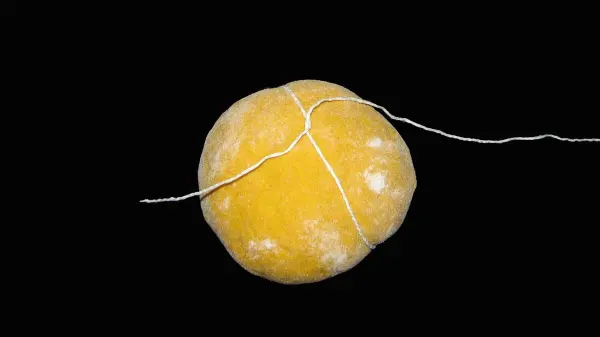 Ein leuchtend gelbes Kürbisbrötchen liegt auf einer dunklen Fläche und wird mit weißem Bindfaden zusammengehalten.