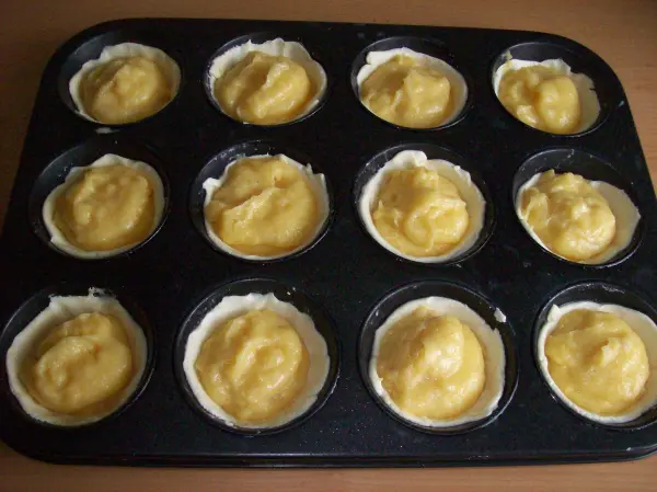 In einer Muffinform liegen zwölf gefüllte Teigschalen, bereit zum Backen von Pasteis de Belem, in einer hellen Umgebung.