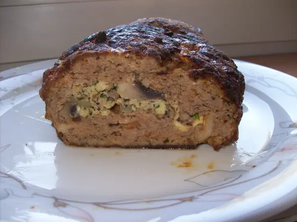 Ein gefüllter falscher Hase mit einer goldbraunen Kruste liegt auf einem weißen Teller, mit Kräutern, Champignons und Ei sichtbar.