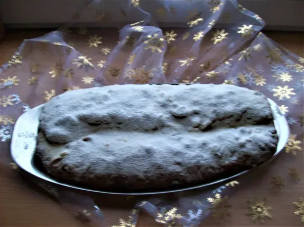 Ein Christstollen mit Puderzucker liegt auf einem silbernen Teller, umgeben von einem glitzernden, goldenen Tuch.