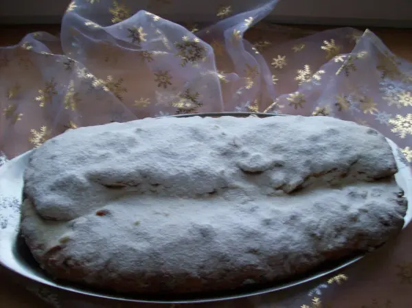 Ein Christstollen mit Mehlstaub liegt auf einem silbernen Teller, umgeben von glitzerndem Weihnachtsdekor im Hintergrund.