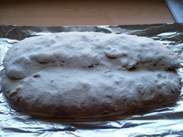 Ein Christstollen mit einer Puderzucker-Schicht liegt auf Alufolie, umgeben von einer einfachen, hellen Tischoberfläche.