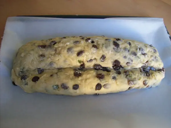 Ein Christstollen mit Marzipan liegt auf einem Backblech, bereit zum Backen, umgeben von einer neutralen Arbeitsfläche.