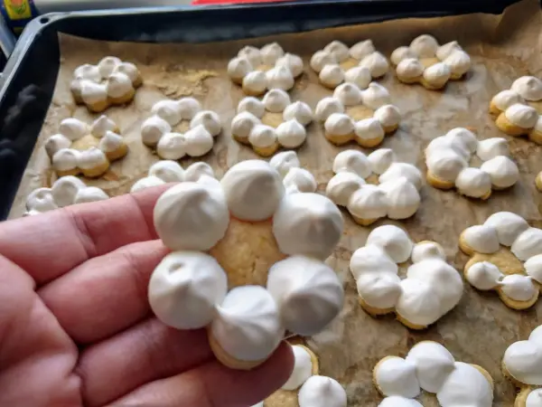 Hand hält ein Gebäckstück in Form eines Kranzes aus Meringue, umgeben von mehreren weiteren Meringen auf einem Backblech.