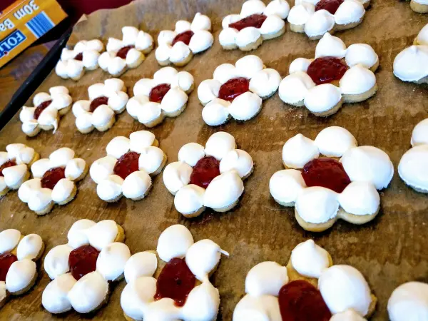 Die Kekse in Form von Blumen liegen auf einem Backblech und sind mit roter Marmelade gefüllt. Der Hintergrund zeigt eine Küche.