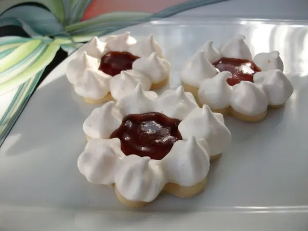 Drei nussige Plätzchen mit einer glänzenden Marmeladenfüllung liegen auf einem weißen Teller mit floralen Mustern.