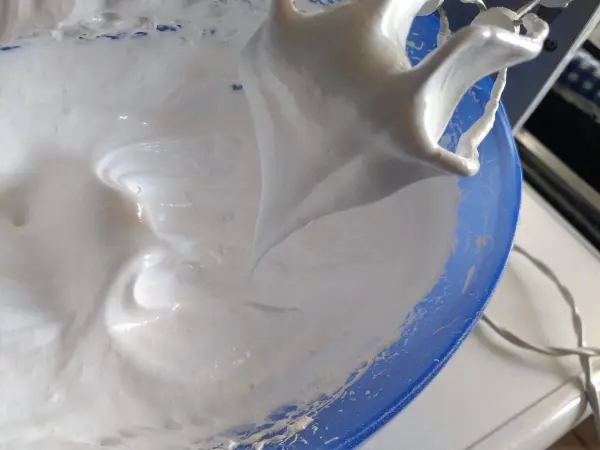 Ein elektrischer Mixer schwingt über eine Schüssel mit fluffigem Eischnee, während die Küchenumgebung unscharf im Hintergrund bleibt.
