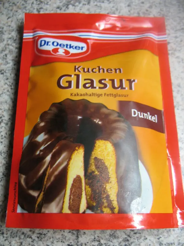 Ein dunkelglasierter Kuchen ist in der Mitte durchgeschnitten, zeigt ein marmoriertes Muster und liegt vor einer roten Packung.