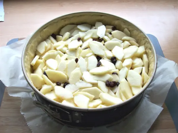 Ein Käse-Apfel-Kuchen befindet sich in einer Springform, gefüllt mit dünn geschnittenen Äpfeln und Rosinen.
