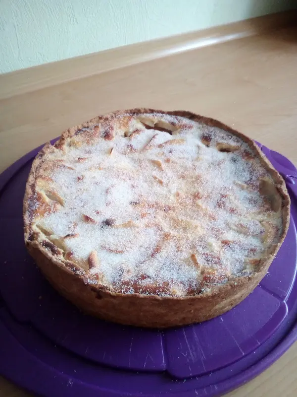 Ein Käse-Apfel-Kuchen steht frisch gebacken auf einem lila Tortenteller, mit Zuckerglasur und einer goldbraunen Kruste.