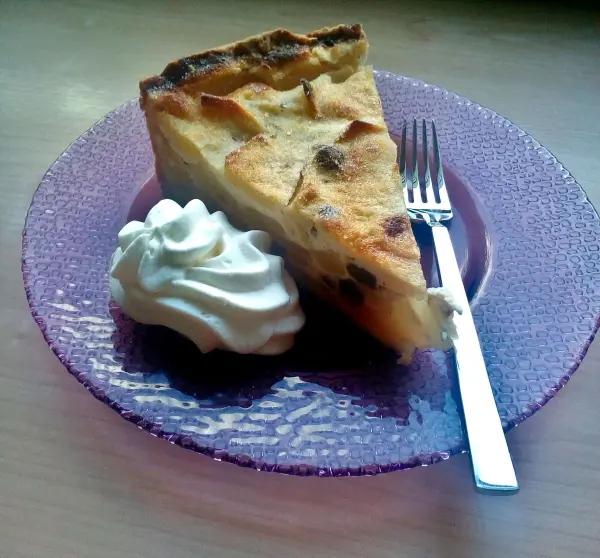Ein Stück Käse-Apfel-Kuchen liegt auf einem lila Teller, neben einem Klecks Sahne und einer Gabel.