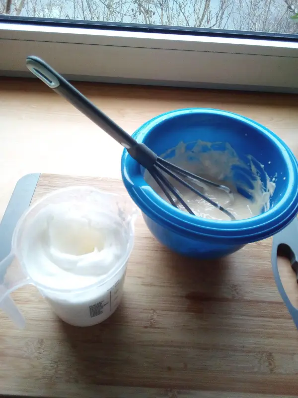 Eine blaue Schüssel mit einer Mischung steht neben einem Messbecher mit Sahne, beide auf einer Holzoberfläche beim Backen.