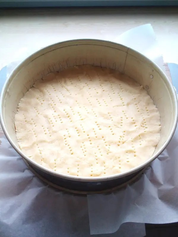 Ein roher Teigboden für Käse-Apfel-Kuchen ruht in einer Springform, umgeben von Backpapier auf einem Holzbrett.