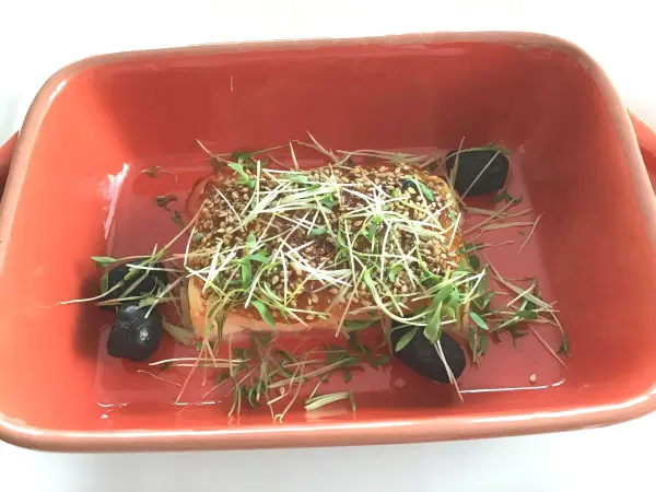 Gebackener Feta liegt in einer rot-orangefarbenen Auflaufform, garniert mit Sesam und frischen Kräutern, umgeben von dunklen Beilagen.