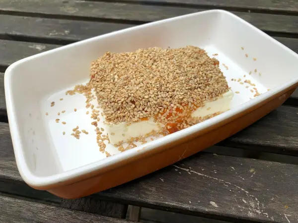 Gebackener Feta liegt in einer weißen Auflaufform, bedeckt mit Sesam und umgeben von einem Holzuntergrund.