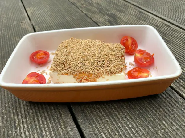 Gebackener Feta liegt in einer Schale, umgeben von Sesam und halbierten Tomaten, auf leicht verwittertem Holz.