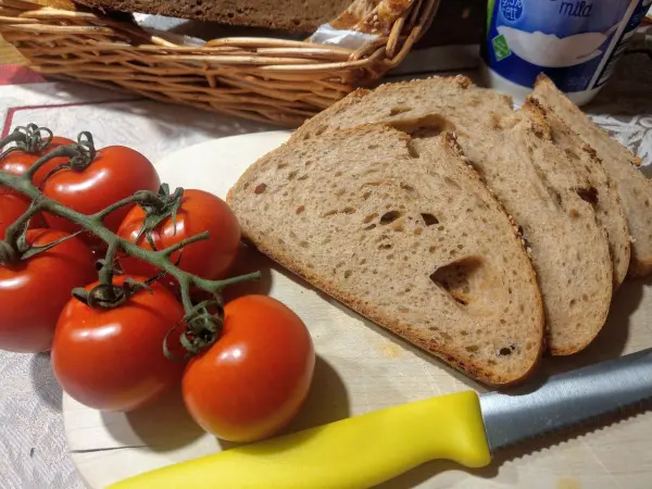 Auf einem Holzbrett liegen Scheiben von Vollkornbrot neben einer Traube von frischen Tomaten, während ein gelbes Messer bereitliegt.