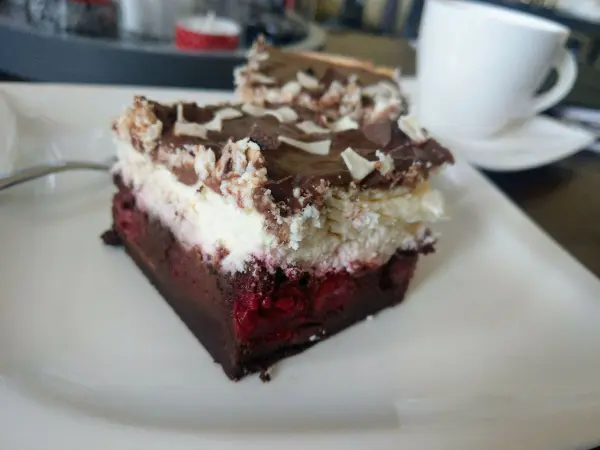 Ein Stück Schwarzwälder Käsekuchen liegt auf einem weißen Teller, umgeben von einer Tasse Kaffee auf einem Tisch.