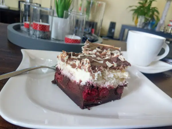 Ein Stück Schwarzwälder Käsekuchen mit Schokolade und Kirschen liegt auf einem weißen Teller in einer gemütlichen Umgebung.