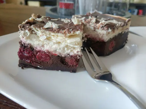 Ein Stück Schwarzwälder Käsekuchen liegt auf einem weißen Teller, garniert mit Schokolade und Kirschen, neben einer Gabel.