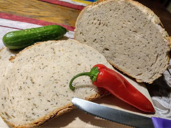 Ein golden-braunes Mohn-Dinkel-Brot liegt auf einem Holzbrett, umgeben von einer grünen Gurke und einer roten Chili.