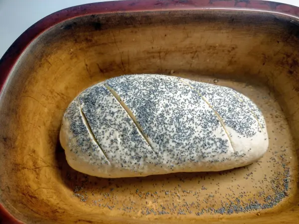 Ein Laib Mohn-Dinkel-Brot ruht in einer Holzform und wartet auf das Backen, mit gesprenkelter Mohnsaat auf der Oberfläche.