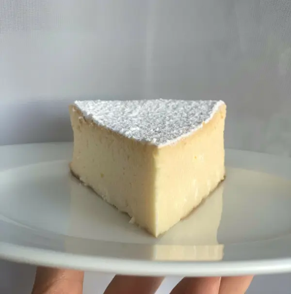 Ein Stück saftiger Käsekuchen ohne Boden liegt auf einem weißen Teller, präsentiert in natürlichem Licht.