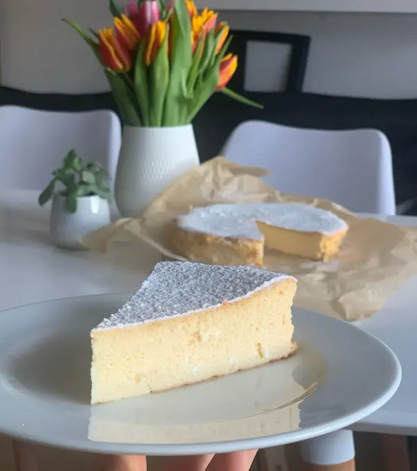 Ein Stück Käsekuchen ohne Boden steht auf einem Teller, umgeben von frischen Blumen und einem weiteren Kuchen im Hintergrund.