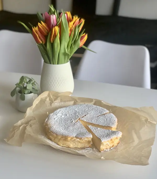 Ein saftiger Käsekuchen ohne Boden liegt auf Backpapier, während eine Scheibe herausgeschnitten ist, umgeben von Tulpen.