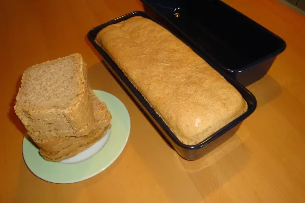 Ein dunkles Dinkelbrot liegt in einer Kastenform, während mehrere Scheiben auf einem Teller daneben gestapelt sind.