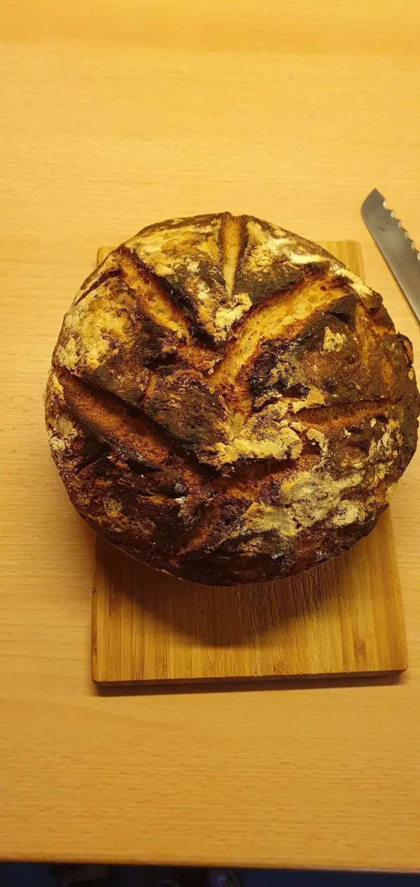 Ein frisch gebackenes Dinkel-Sauerteigbrot ruht auf einem Holzbrett, daneben liegt ein gezacktes Brotmesser.