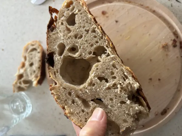 Ein Stück Dinkel-Sauerteigbrot wird auf einer Küchenplatte gehalten, daneben ein rundes Holzbrett mit Brotkrümeln.