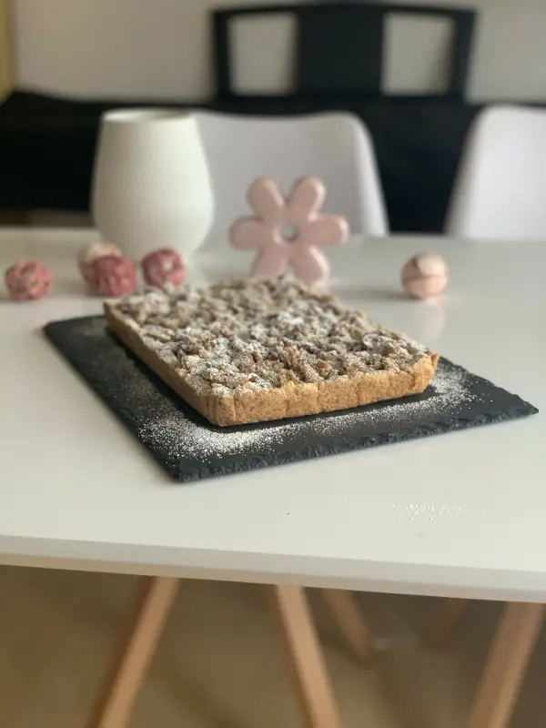Ein Apfelstreuselkuchen mit Mandeln liegt auf einem schwarzen Teller, umgeben von einer weißen Vase und Dekoration.