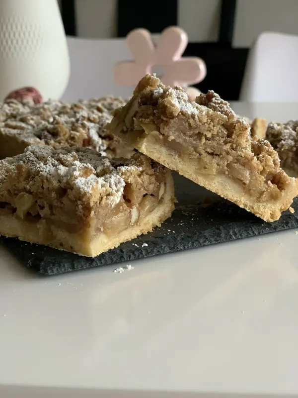 Ein knuspriger Apfelstreuselkuchen mit Mandeln liegt auf einem dunklen Stein, umgeben von hellen Farben und einem stilvollen Hintergrund.