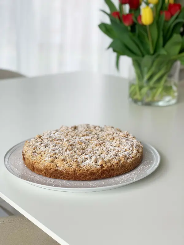 Ein Apfelstreuselkuchen mit Mandeln liegt auf einem durchsichtigen Teller auf einem weißen Tisch, umgeben von frischen Blumen.