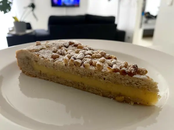 Ein Stück Torta della Nonna liegt auf einem weißen Teller, umgeben von einem modernen Wohnraum mit einem Sofa im Hintergrund.