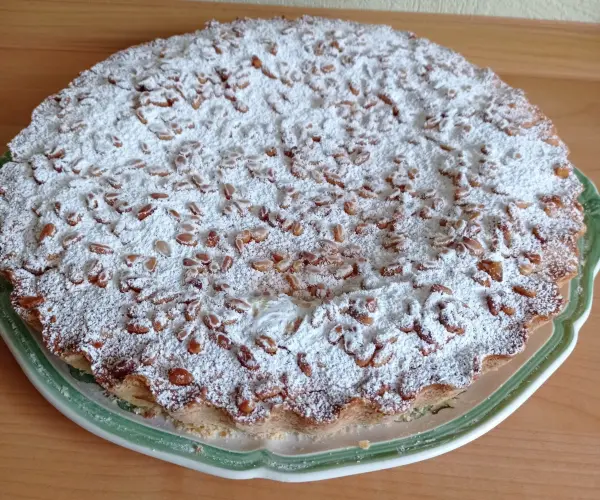 Eine Torta della Nonna liegt auf einem grünen Teller, bestreut mit Puderzucker und Mandeln, auf einem Holztisch.